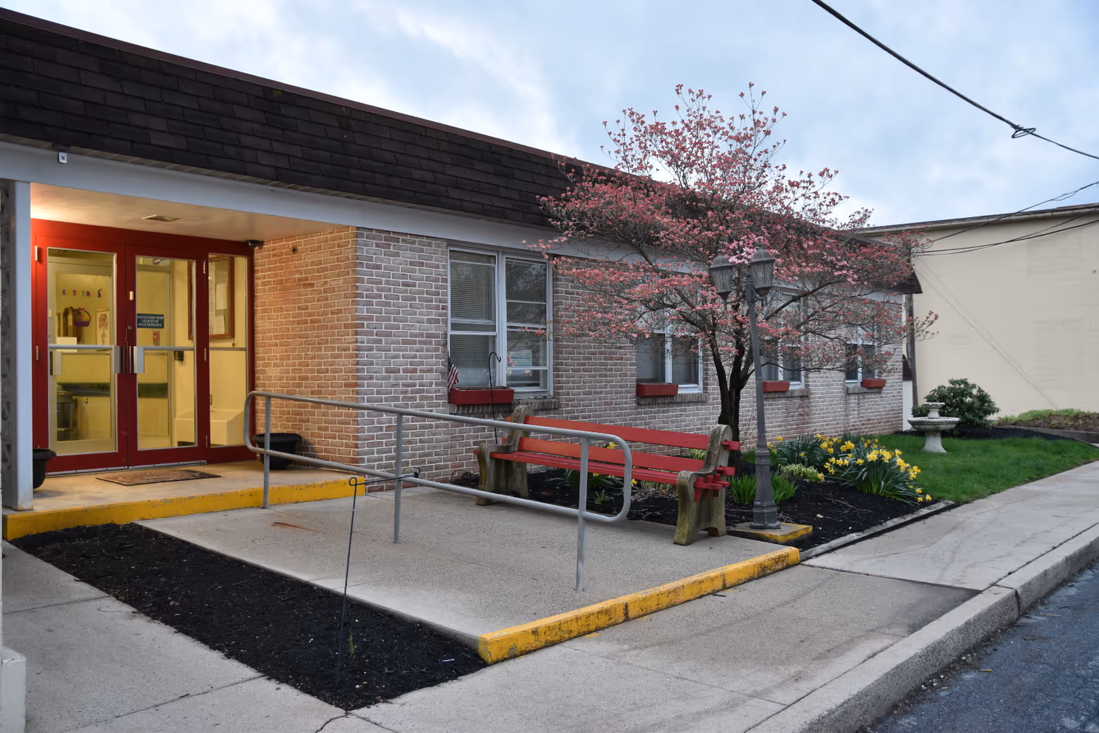 Entrance to a brick building with red double doors, a wheelchair ramp, a red bench, a flowering tree, and a small garden area with yellow flowers and a birdbath.