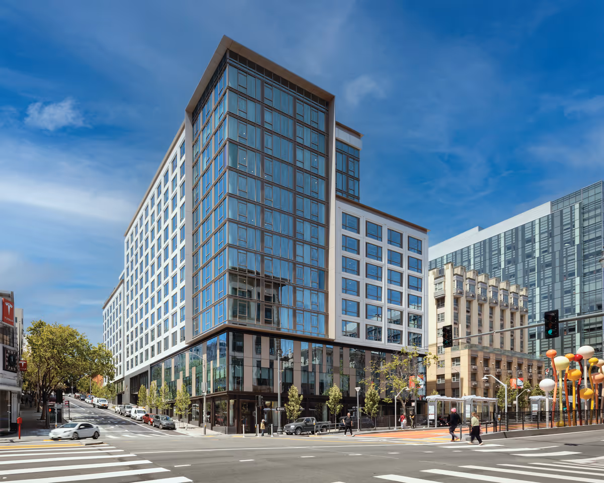 Modern multi-story glass and white-facade building on a city corner with pedestrians, cars, and traffic lights under a blue sky.