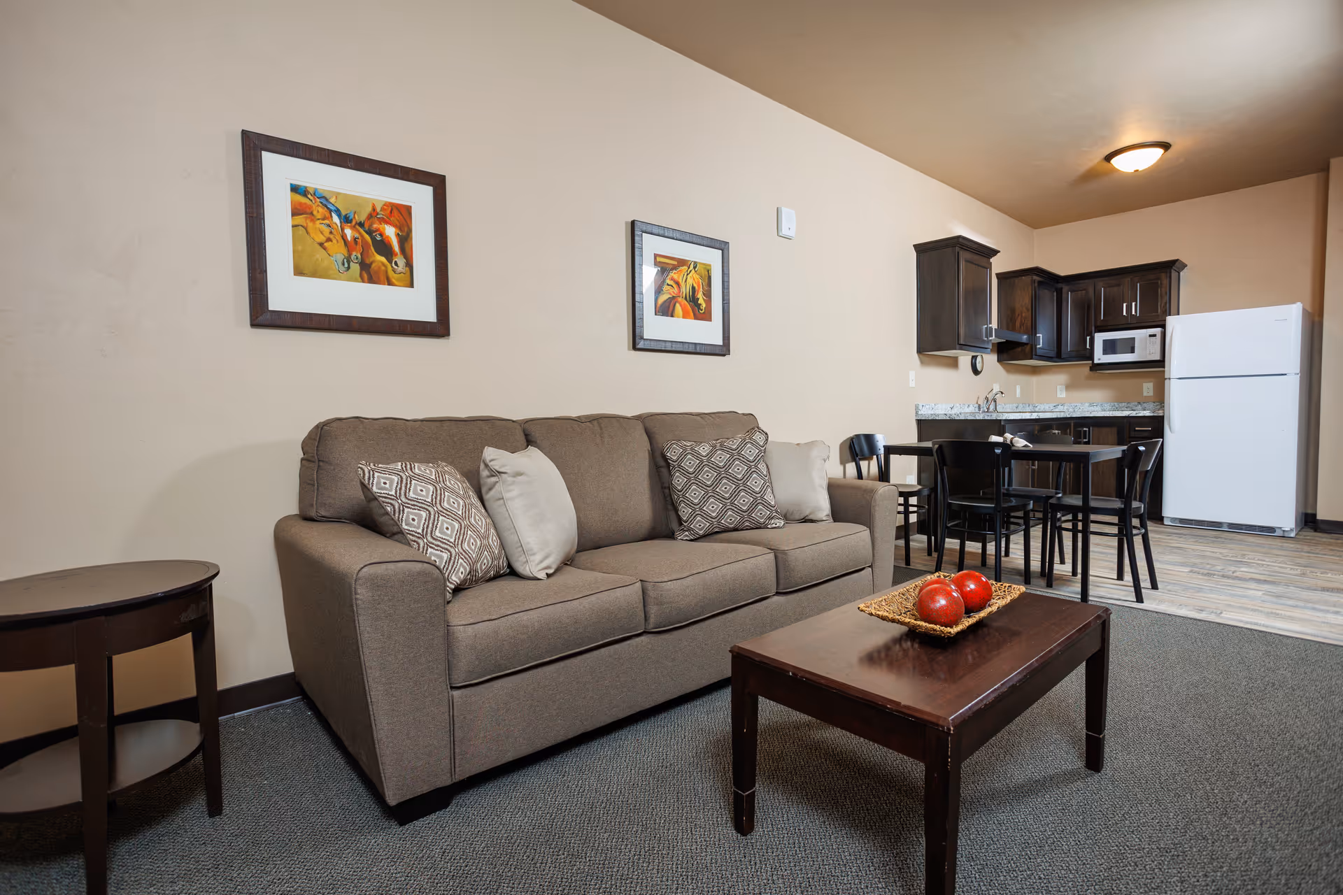 Open-plan living area with a brown sofa and coffee table in front of wall art, and a kitchenette with dining table and refrigerator in the background.