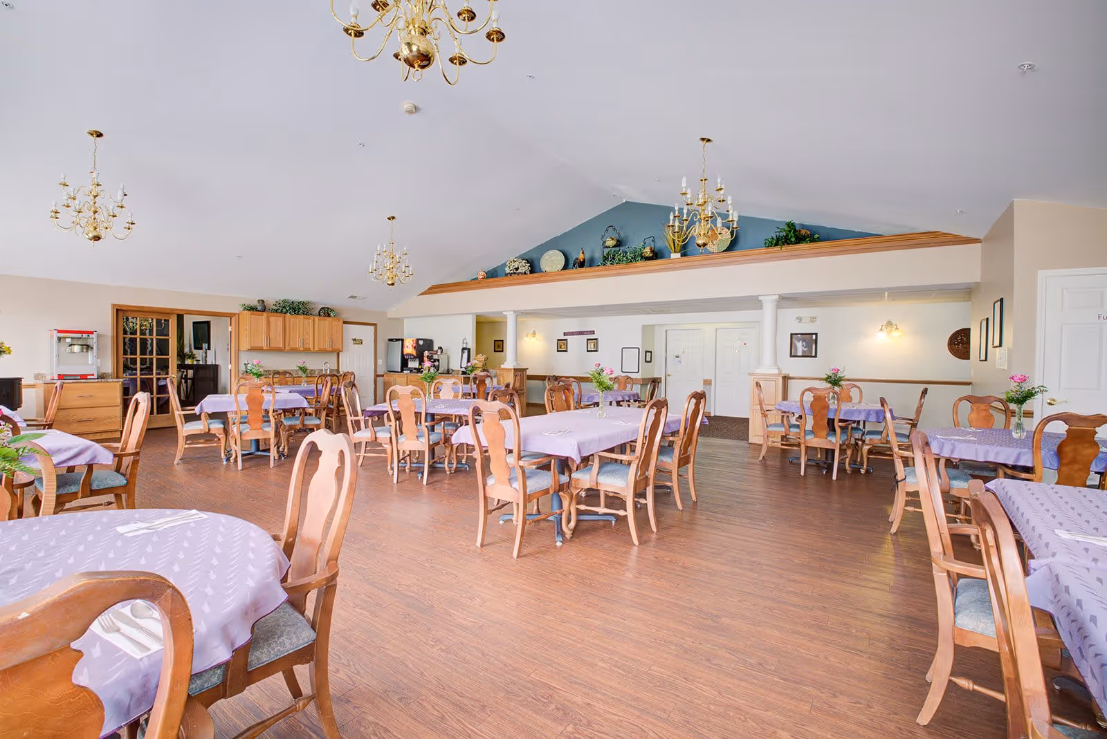 Spacious dining room with multiple wooden tables and chairs covered with lavender tablecloths. The room has wooden flooring, several chandeliers hanging from a high ceiling, and decorative plants and items on a ledge above a partial wall. There is a popcorn machine and a beverage dispenser in the back corner.