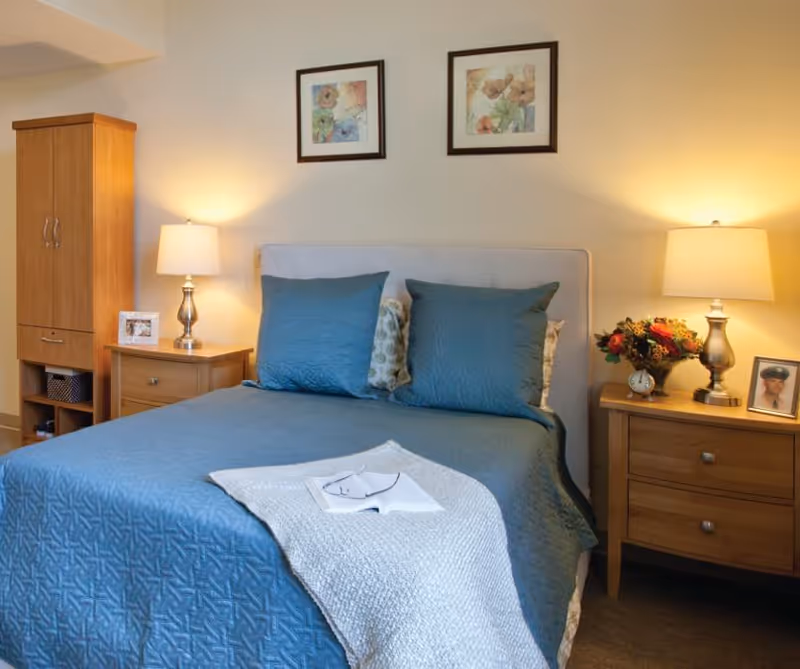 A neatly made bedroom with a blue quilted bed, two bedside tables with lamps and decor, and framed artwork above the headboard.