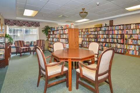 A cozy library room with a round wooden table surrounded by four cushioned chairs in the center. The walls are lined with bookshelves filled with books. Two armchairs with striped upholstery are placed near a window with floral valances, and there are plants on the windowsill and a small table. The ceiling has recessed lighting and a decorative light fixture.