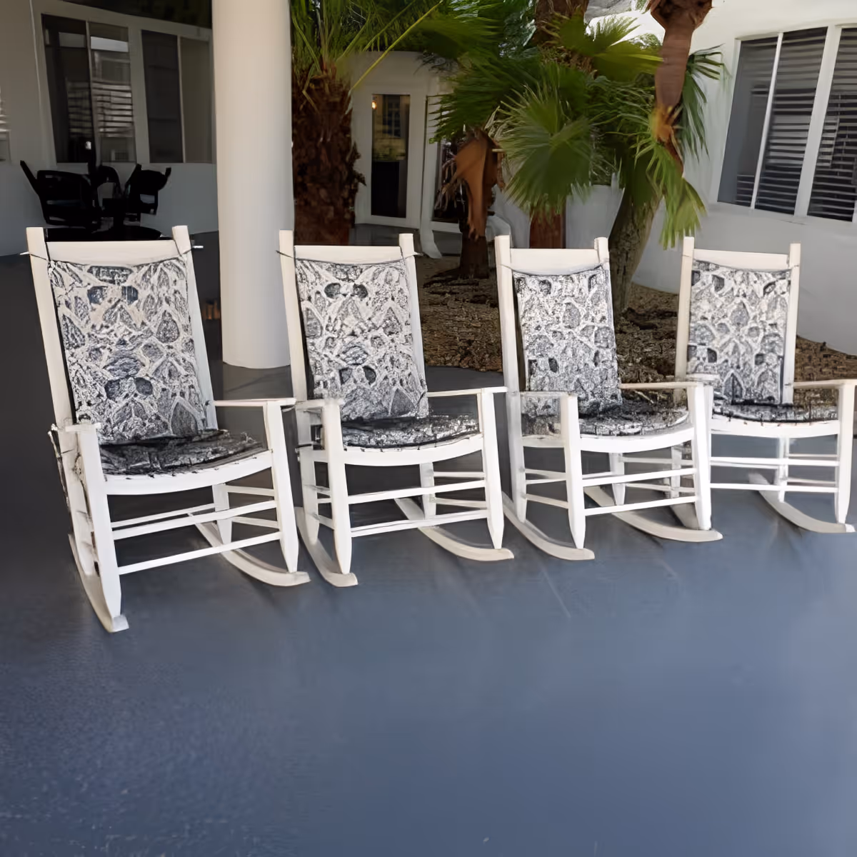 Four white wooden rocking chairs with patterned cushions arranged in a row on a covered patio area with a gray floor. Behind the chairs are white columns, windows, and green palm plants.