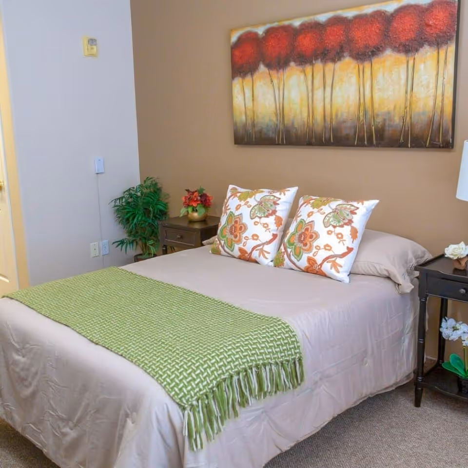 A cozy bedroom with a bed dressed in neutral linens, two floral pillows, a green throw, bedside tables, and a painting above the headboard.