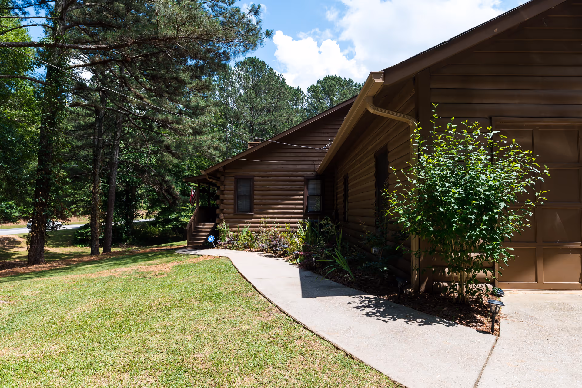Front exterior of a brown wooden cabin-style house with a garage, concrete walkway, lawn, and surrounding trees.