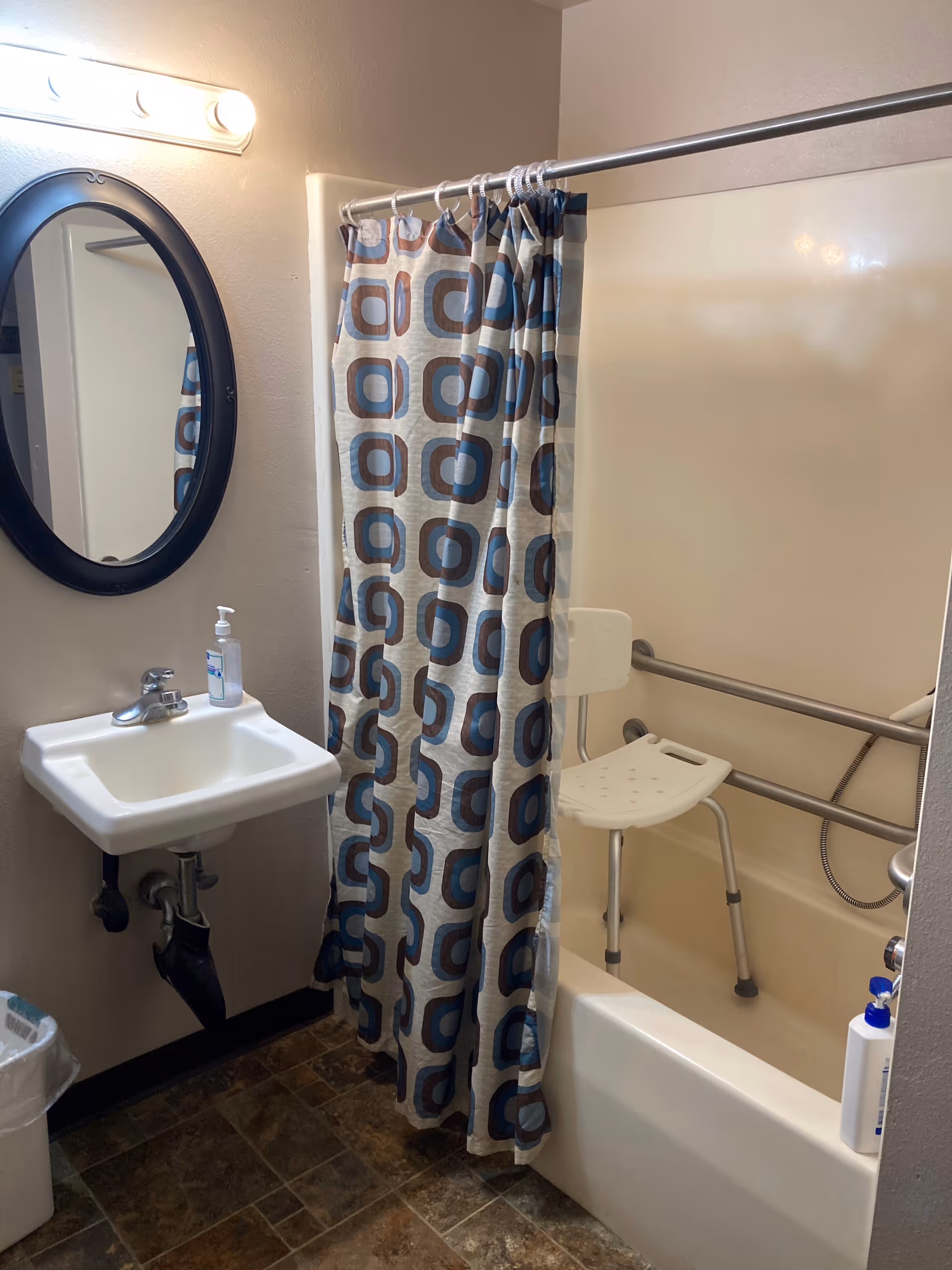 Small accessible bathroom with a sink and oval mirror, patterned shower curtain, bathtub with grab bars and a shower chair.