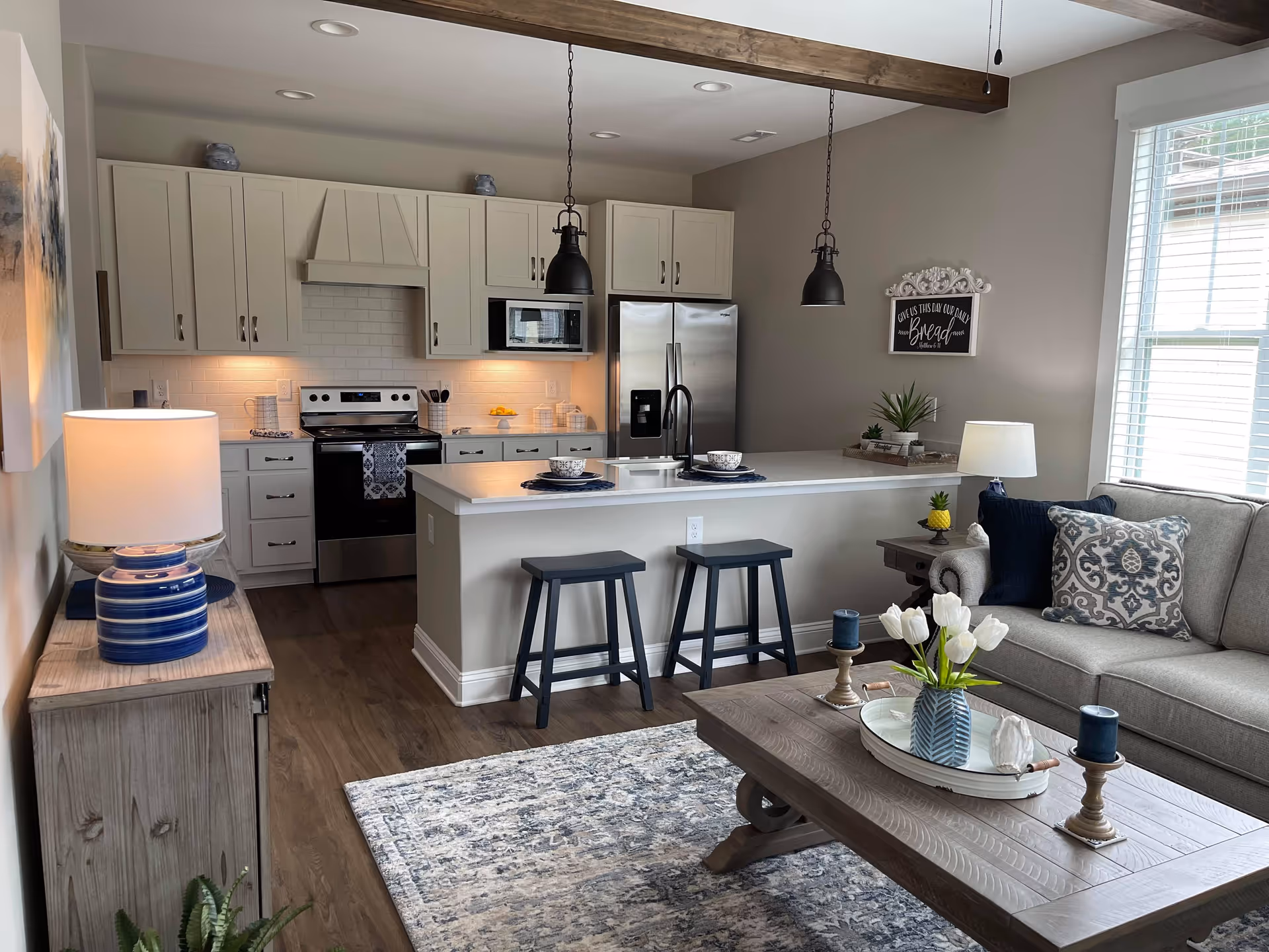 A modern open-concept living area featuring a kitchen with white cabinets, stainless steel appliances, and a large island with two stools. The living room has a gray sofa with decorative pillows, a wooden coffee table with a vase of white tulips, and a side table with a lamp. The room is well-lit with natural light from a window with white blinds.