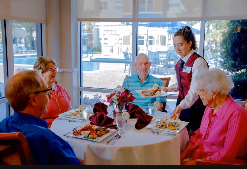 A group of four elderly people sitting around a round dining table with white tablecloth, enjoying a meal served by a young female server in a red vest and white shirt. The table is set with plates of food, glasses of water with lemon slices, and a centerpiece of red and white flowers. Large windows in the background show an outdoor pool area.