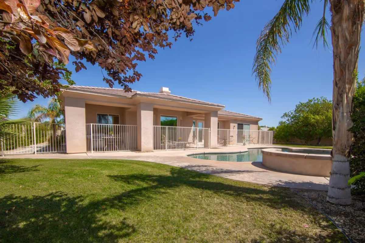 Single-story stucco building with fenced patios and a swimming pool in a grassy yard under a clear blue sky.