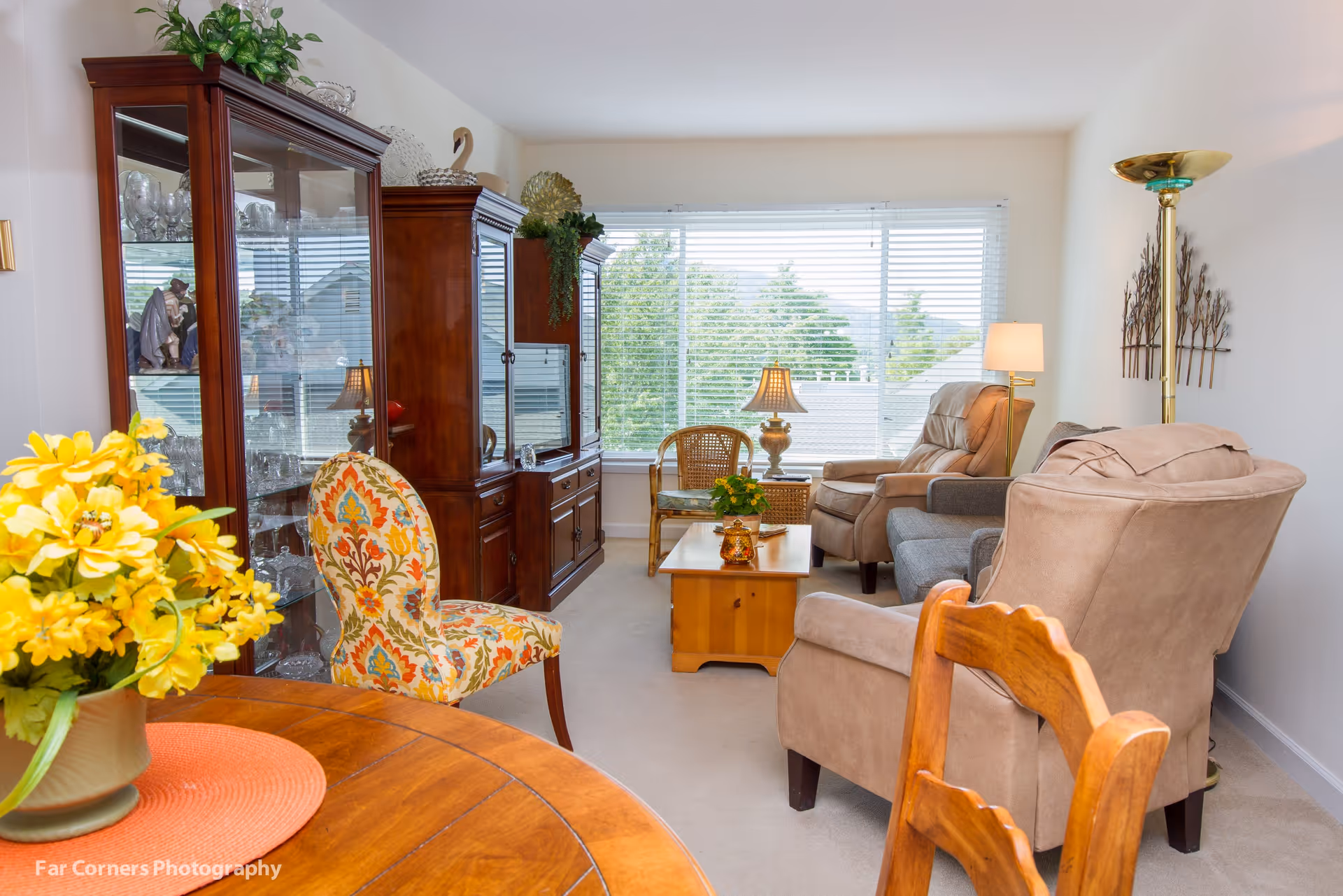 A cozy living room with beige armchairs, a gray sofa, a wooden coffee table with a small plant and decorative items, a wooden china cabinet filled with glassware, a floral patterned chair, a round wooden dining table with a yellow flower arrangement, and large windows with white blinds letting in natural light.
