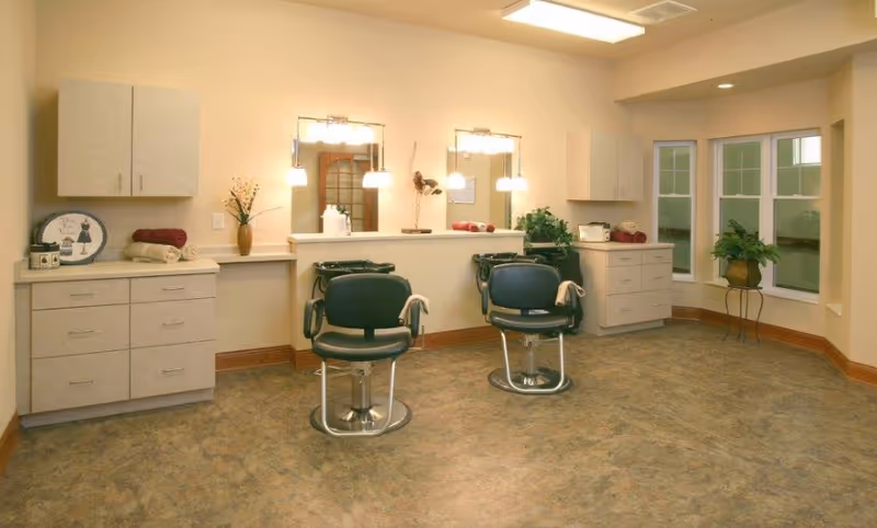 Interior view of a salon area with two black salon chairs in front of mirrors mounted on a half wall. The room has beige walls, cabinets on either side, plants, and soft lighting fixtures above the mirrors. There is a window with multiple panes on the right side.