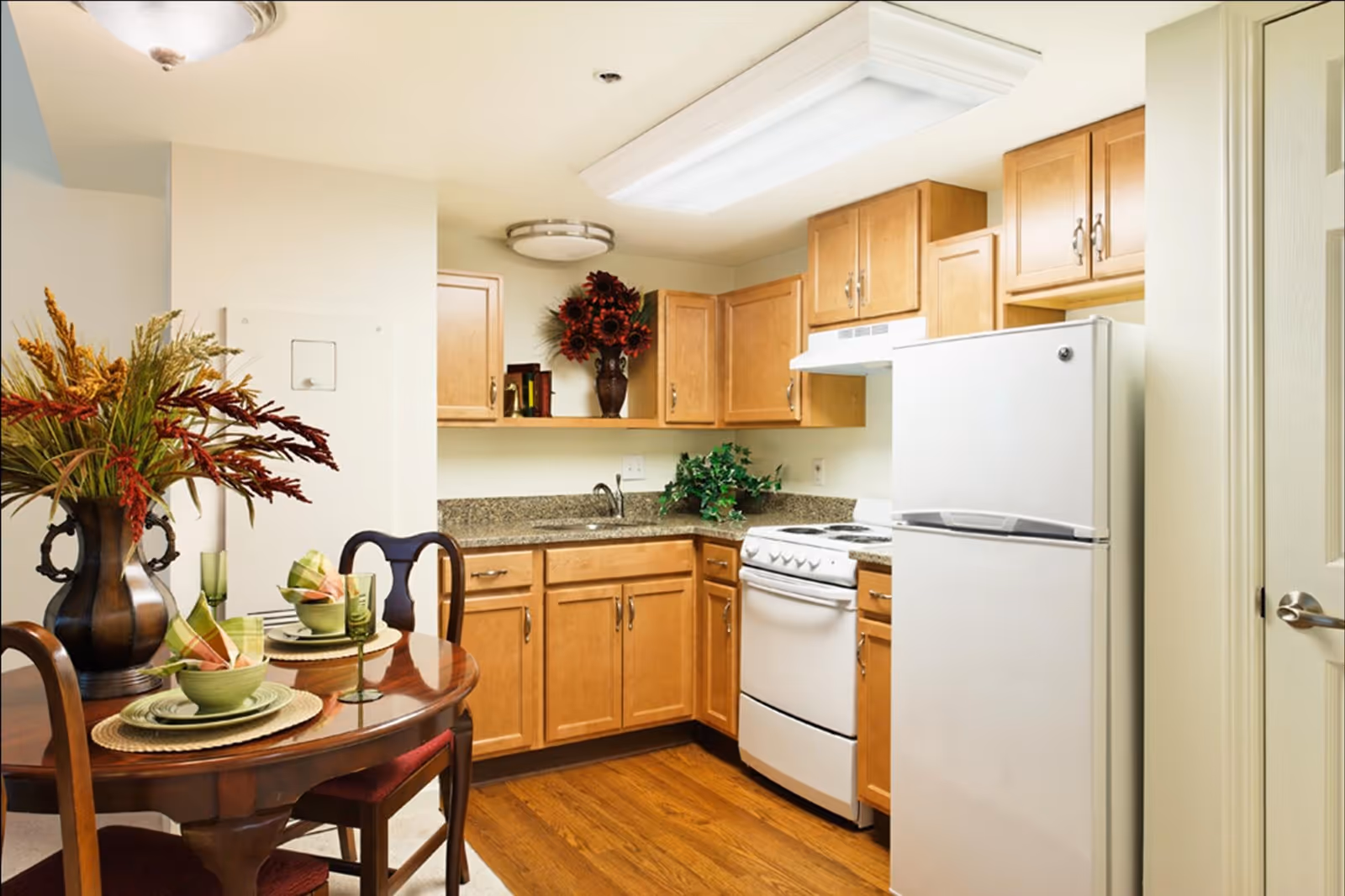 A small kitchen and dining area featuring wooden cabinets, a white refrigerator, a white stove, and a granite countertop. A round wooden dining table is set with green dishes, glasses, and napkins, and decorated with a large vase of flowers. The floor is wooden, and the lighting includes a ceiling fixture and a fluorescent light above the kitchen area.