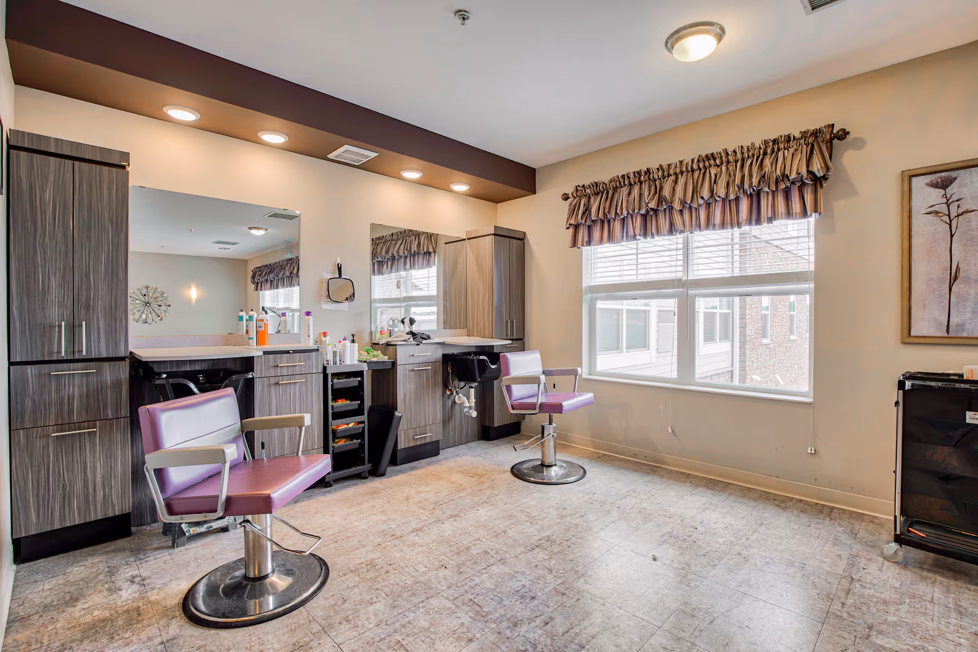 Interior of a salon room with two purple salon chairs, large mirrors, wooden cabinets, and various hair care products on the counter. A window with striped valance curtains lets in natural light, and a framed floral artwork hangs on the wall.