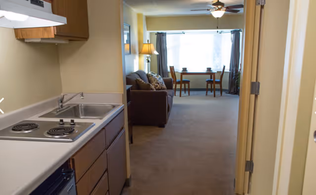 Small studio apartment interior with a kitchenette in the foreground and a living area with a sofa and dining table by a large window.
