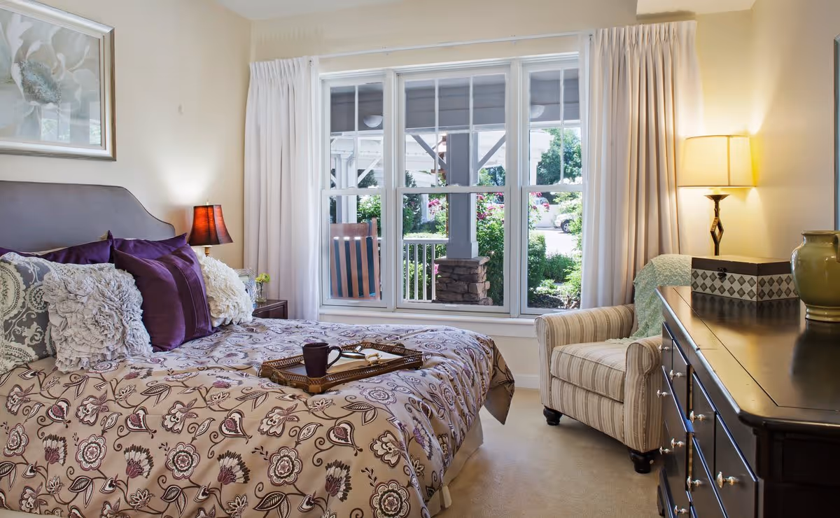 Sunlit bedroom with a patterned bed and decorative pillows, armchair, dresser, and a window looking out to a porch and garden.