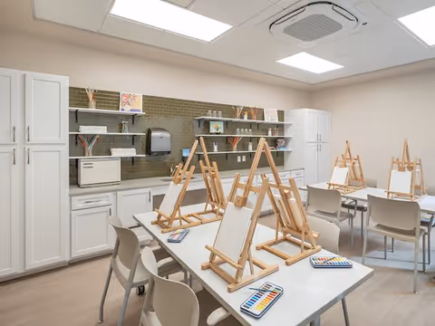 A bright art room with multiple tables and chairs arranged for painting activities. Each table has wooden easels holding blank canvases and watercolor paint sets. The room features white cabinets, open shelves with art supplies, and a green tiled backsplash. The ceiling has recessed lighting and an air conditioning unit.