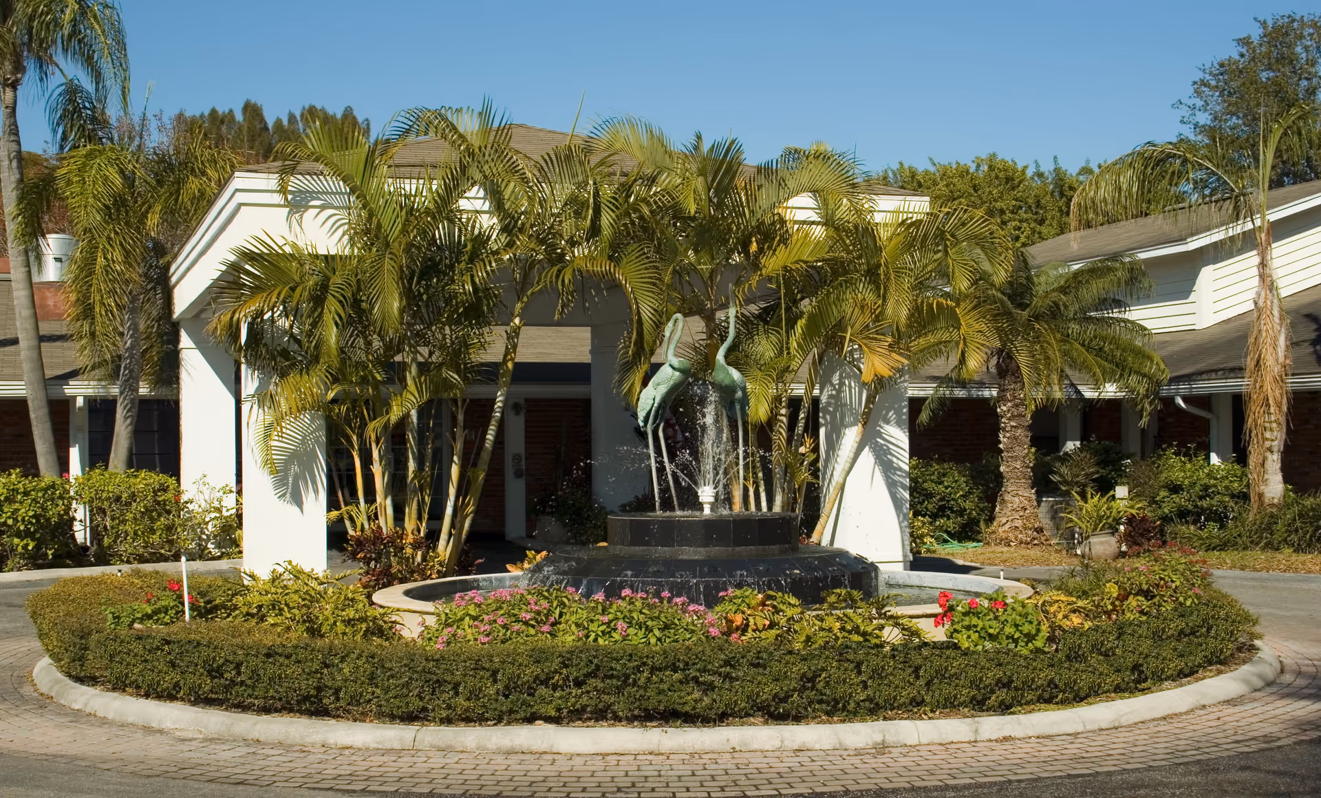 Outdoor circular driveway with a central fountain featuring two crane statues surrounded by palm trees and flowering plants in front of a building entrance at Baytree Lakeside.