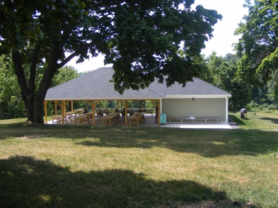 A large open pavilion with a gray roof and wooden support beams situated in a grassy area with large trees providing shade. Under the pavilion are several picnic tables and benches. There is a grill visible on the right side outside the pavilion.