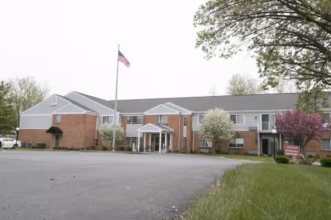 Exterior view of a two-story brick building with white trim, an American flag on a flagpole in front, and several trees with green and purple foliage surrounding the entrance area.