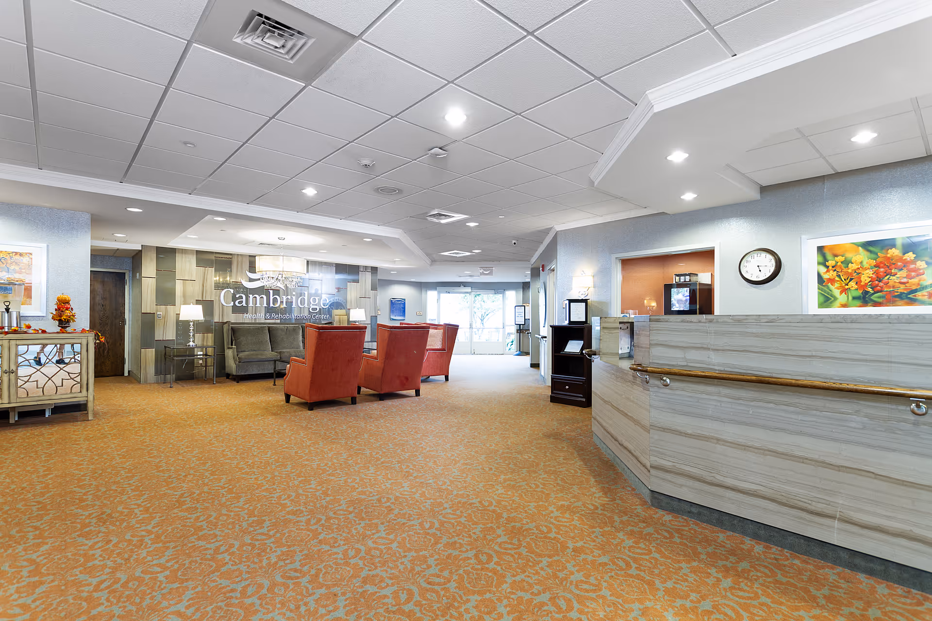 A spacious and well-lit reception area of Cambridge Health and Rehabilitation Center featuring a front desk with a handrail, a seating area with red armchairs and a gray sofa, decorative artwork on the walls, and a patterned carpet floor. The ceiling has recessed lighting and ventilation panels.