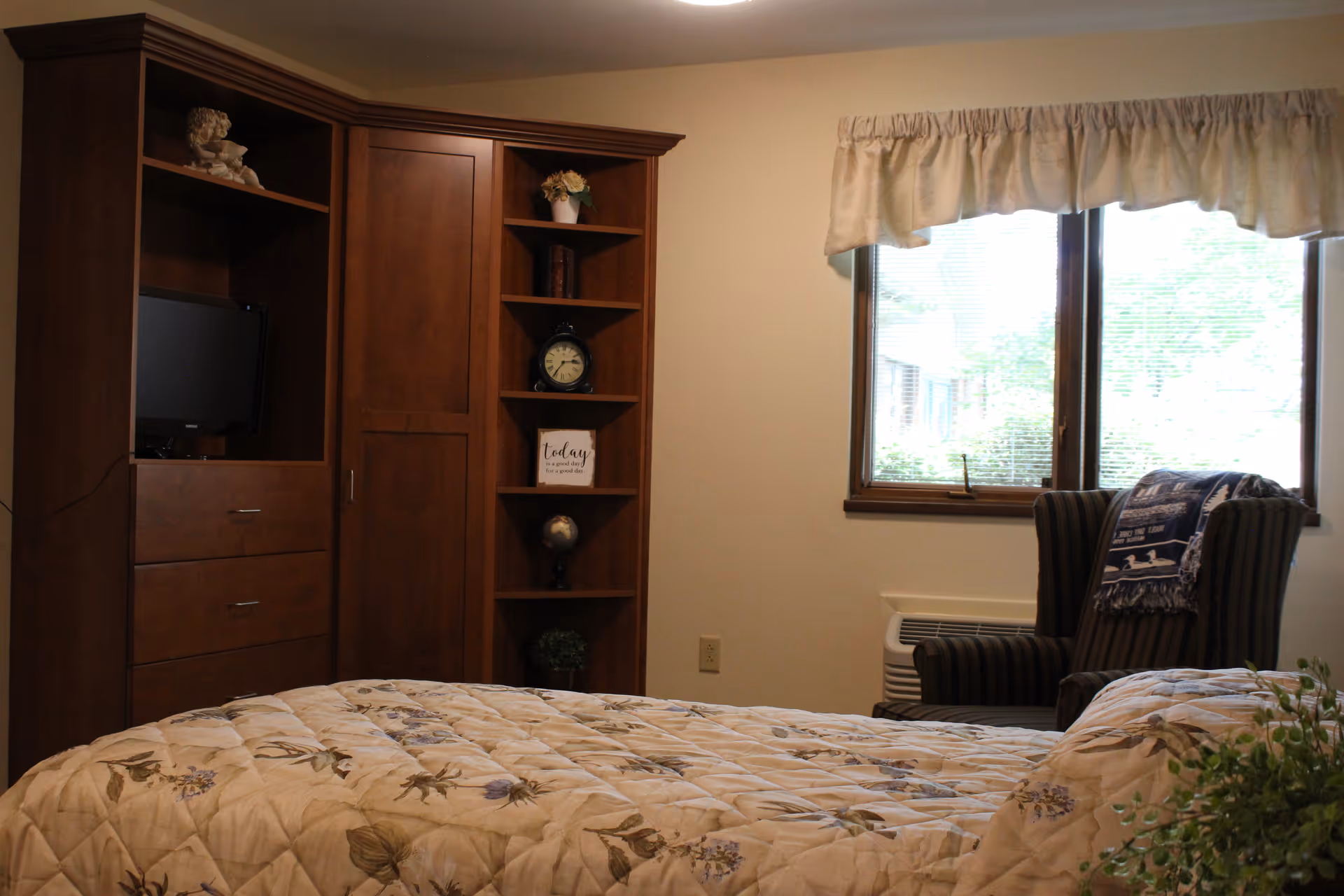 A bedroom with a floral-patterned quilt on the bed, a wooden cabinet with shelves holding decorative items including a clock and a small statue, a window with beige curtains, and a striped armchair with a blanket draped over it.