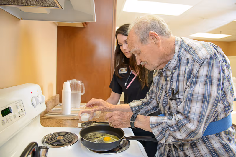 An elderly man cracks an egg into a frying pan on a stove while a caregiver watches in a kitchen.