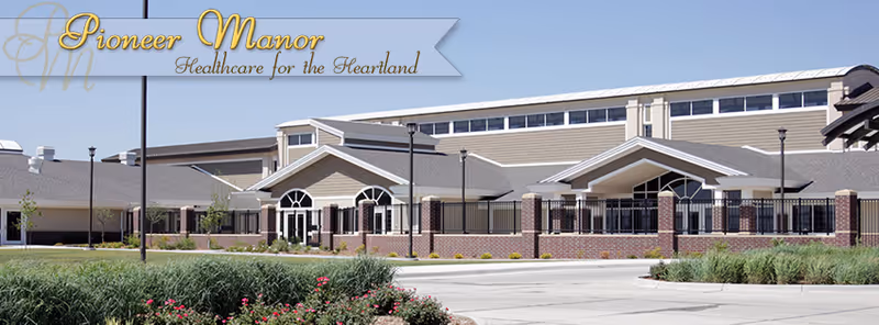 Front exterior of a single-story senior living facility with landscaping and a banner that reads 'Pioneer Manor Healthcare for the Heartland'.