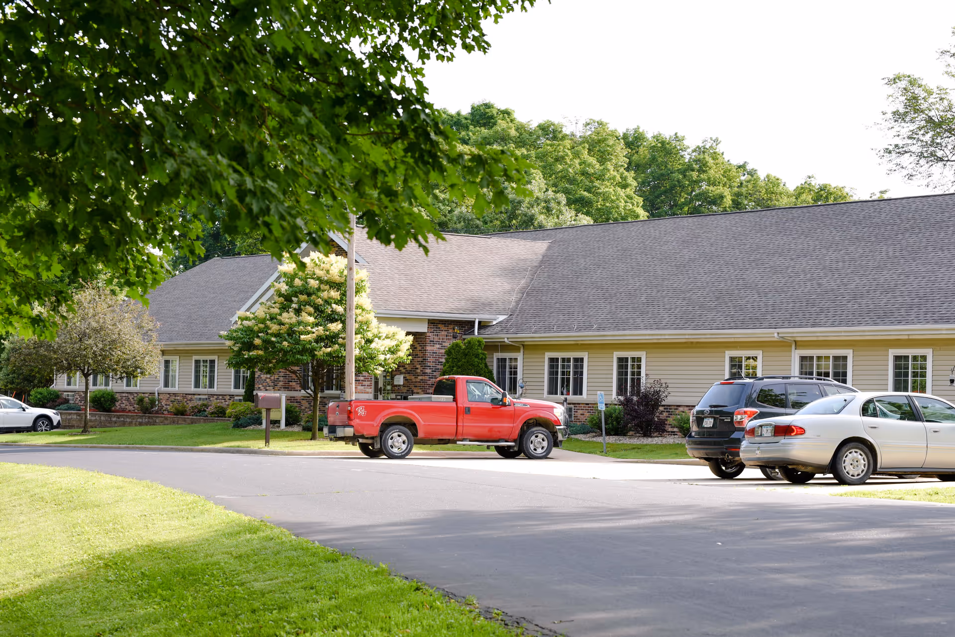 Exterior view of a single-story building with beige siding and brick accents, surrounded by green trees and grass. Several cars, including a red pickup truck, are parked in front of the building along a paved driveway.