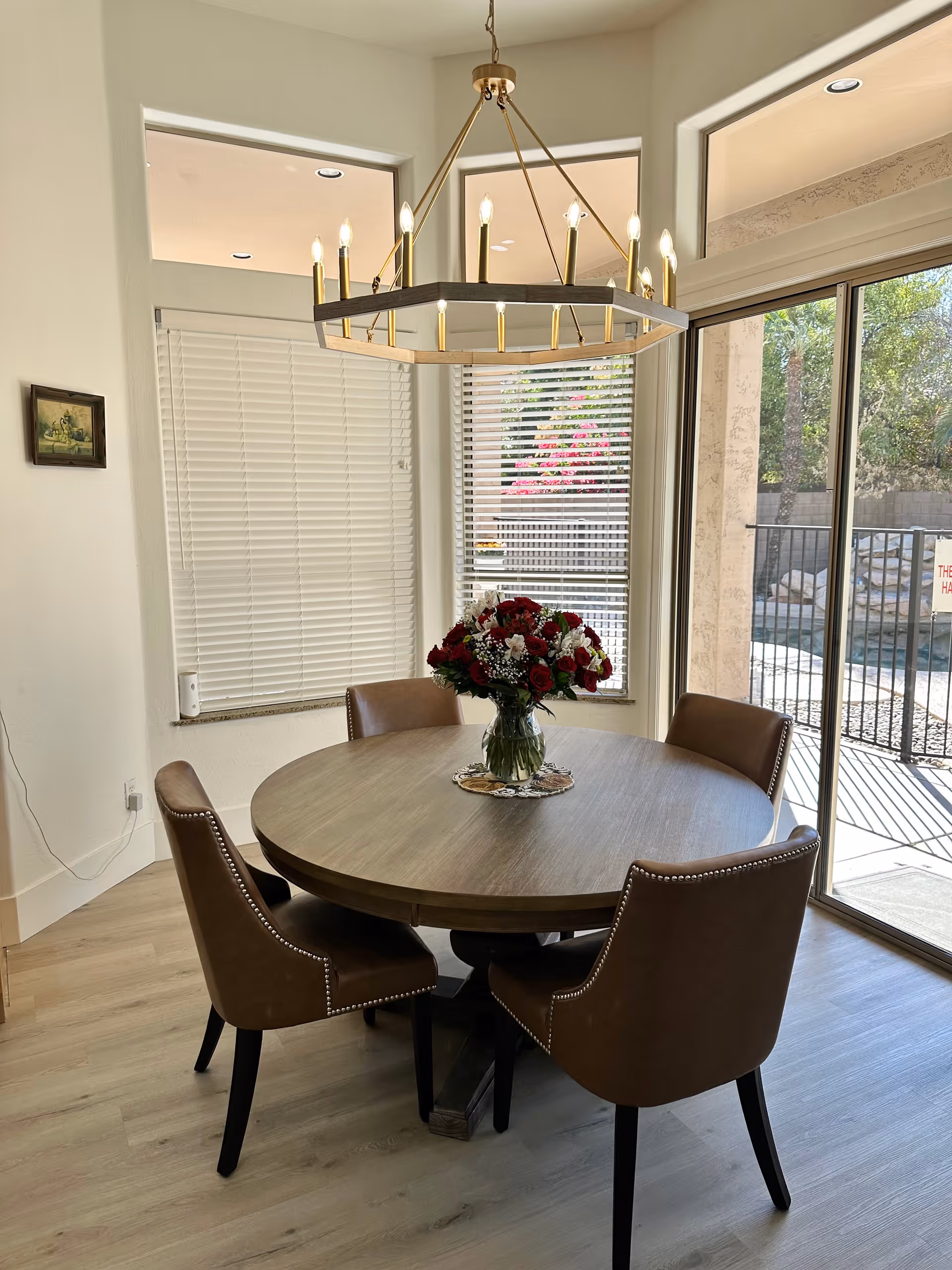 Round wooden dining table with four brown upholstered chairs and a vase of red and white flowers under a chandelier next to sliding glass doors.