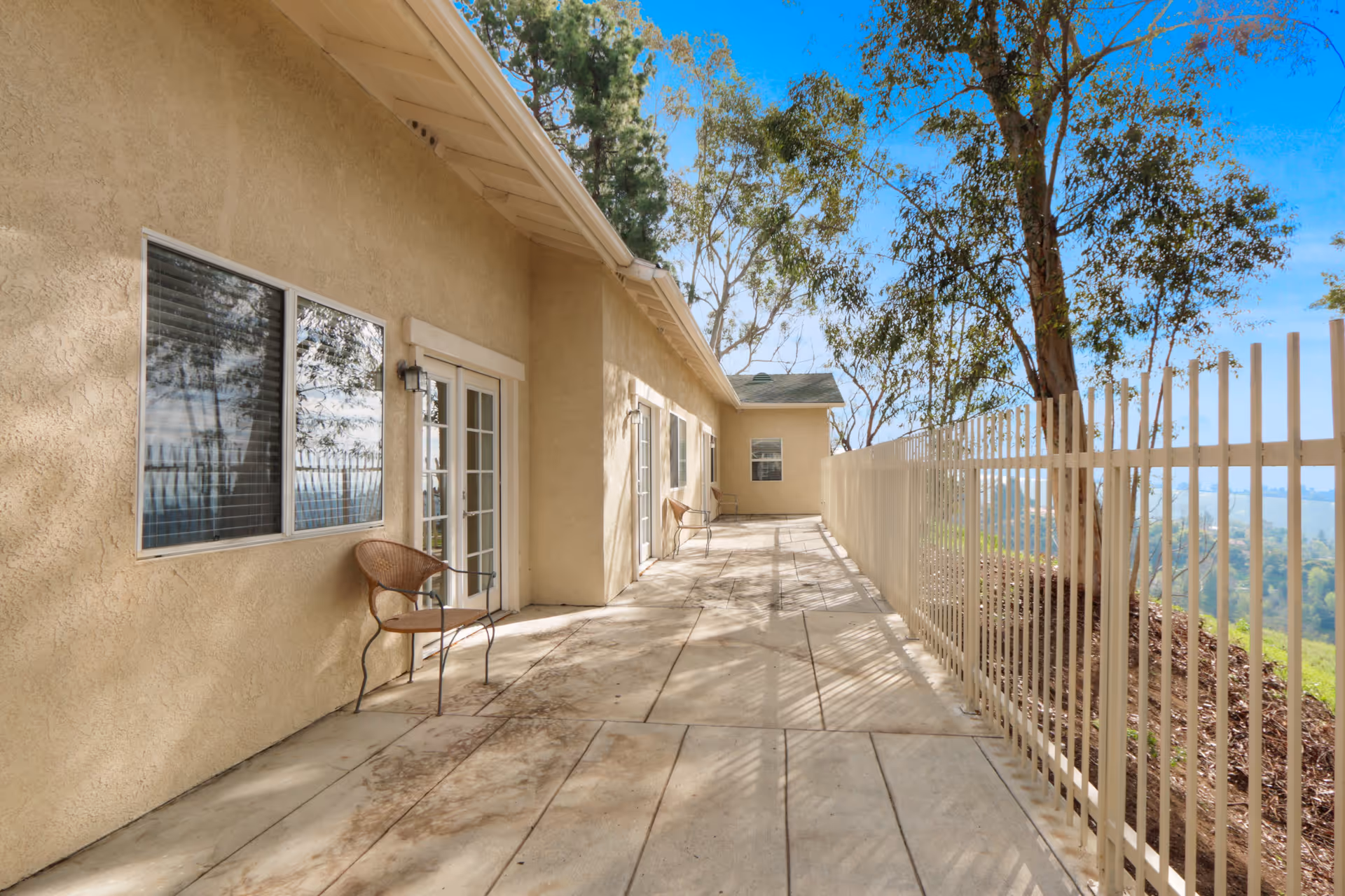 Outdoor patio area with beige stucco walls, multiple windows and glass doors, metal chairs placed along the wall, a white metal fence on the right side, and trees in the background under a clear blue sky.
