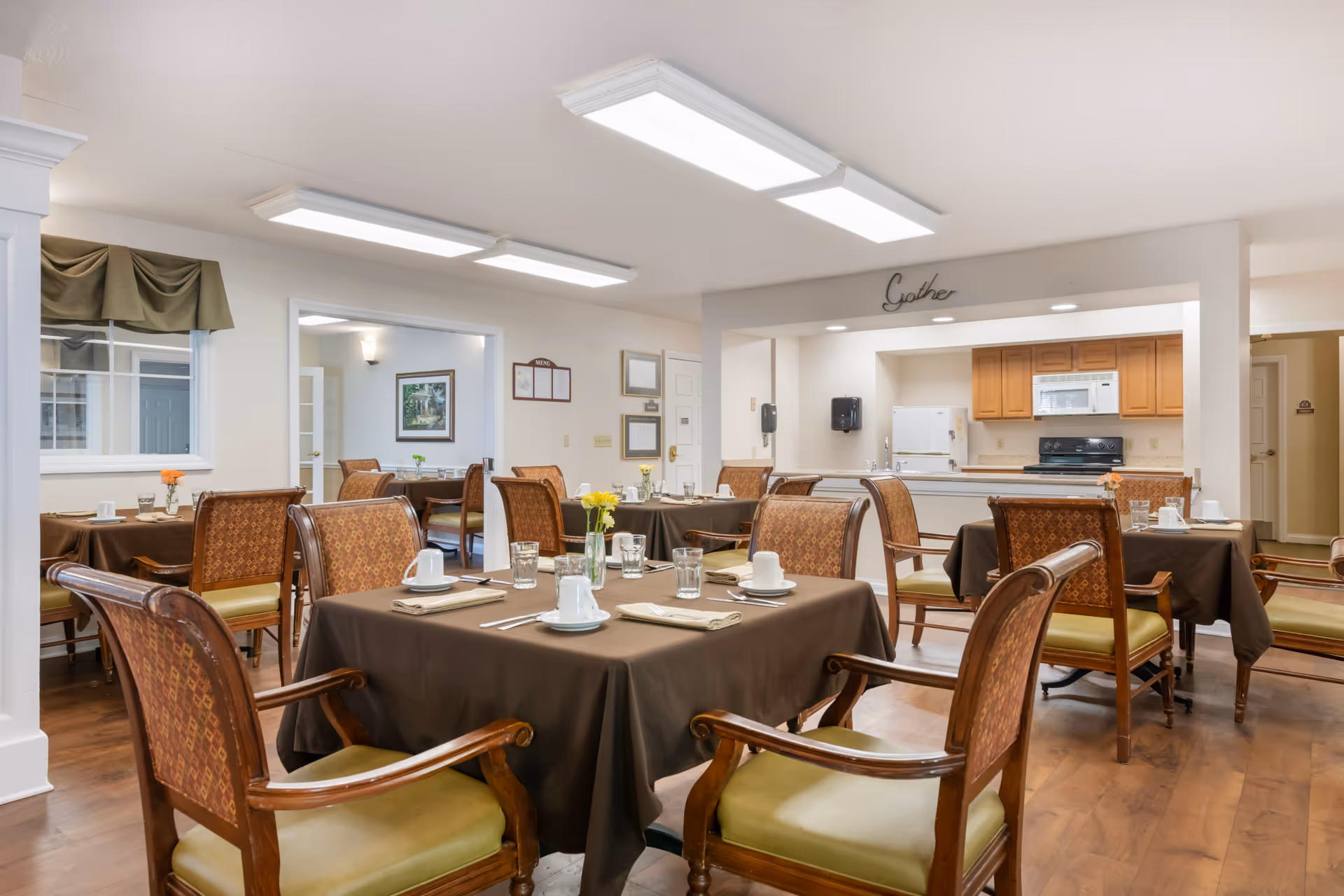 A dining room in a senior living facility with several tables covered in brown tablecloths, each set with white cups, plates, utensils, and glasses. The room has wooden chairs with patterned upholstery and green cushions. In the background, there is a kitchen area with wooden cabinets, a refrigerator, and a stove. The word 'Gather' is displayed above the kitchen pass-through window. The room has bright overhead lighting and hardwood floors.