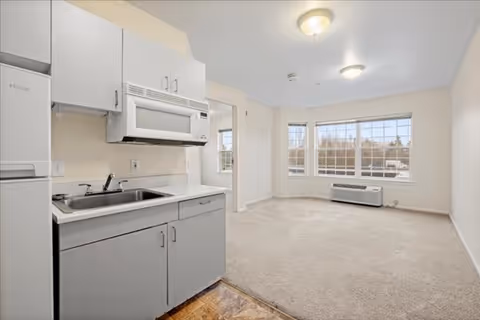 Studio apartment interior with a compact kitchenette opening to a carpeted living area and large bay windows.