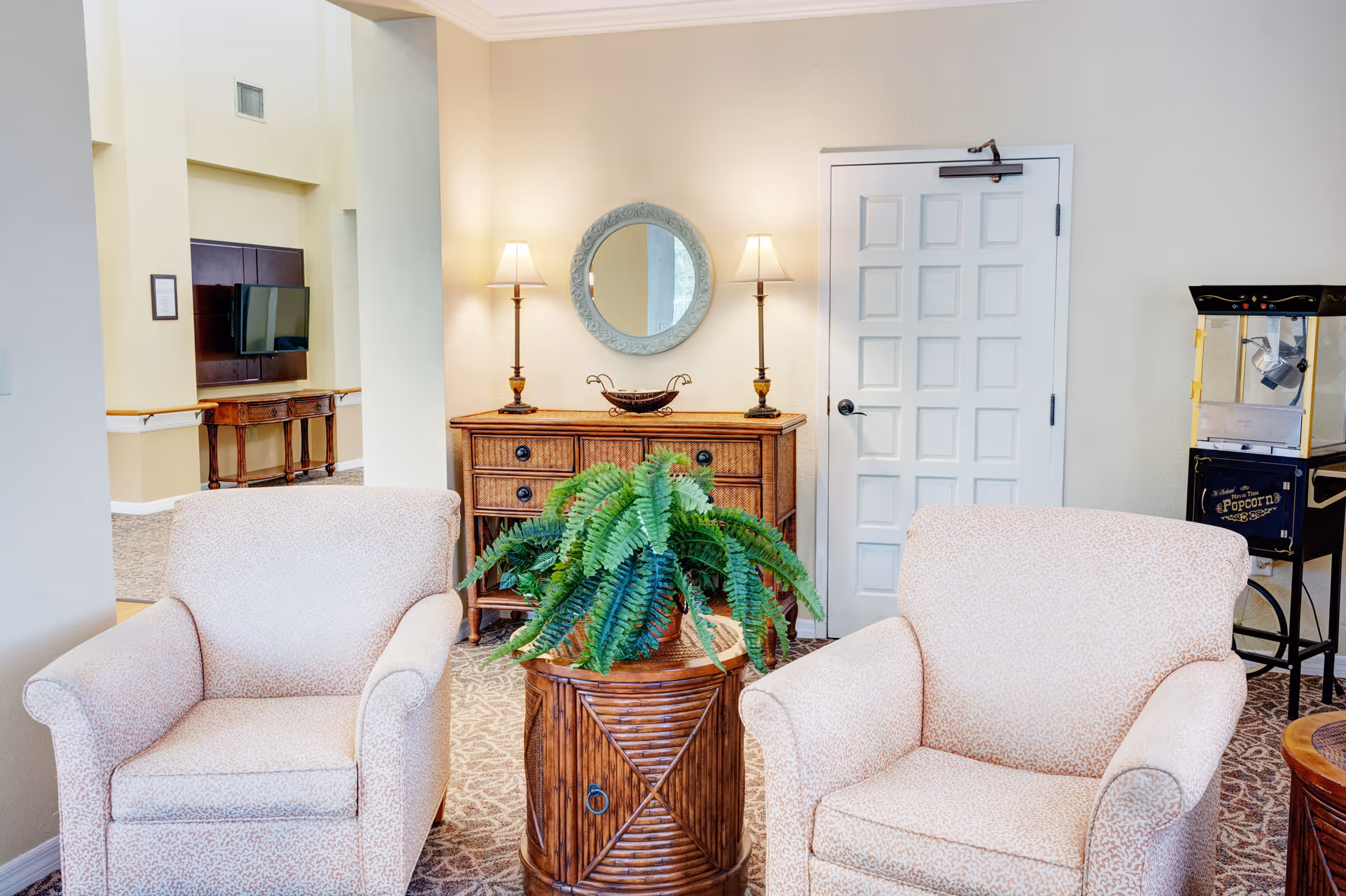 Two upholstered armchairs flank a wooden table with a potted fern in a cozy lounge area with a dresser, round mirror, lamps and a popcorn machine.