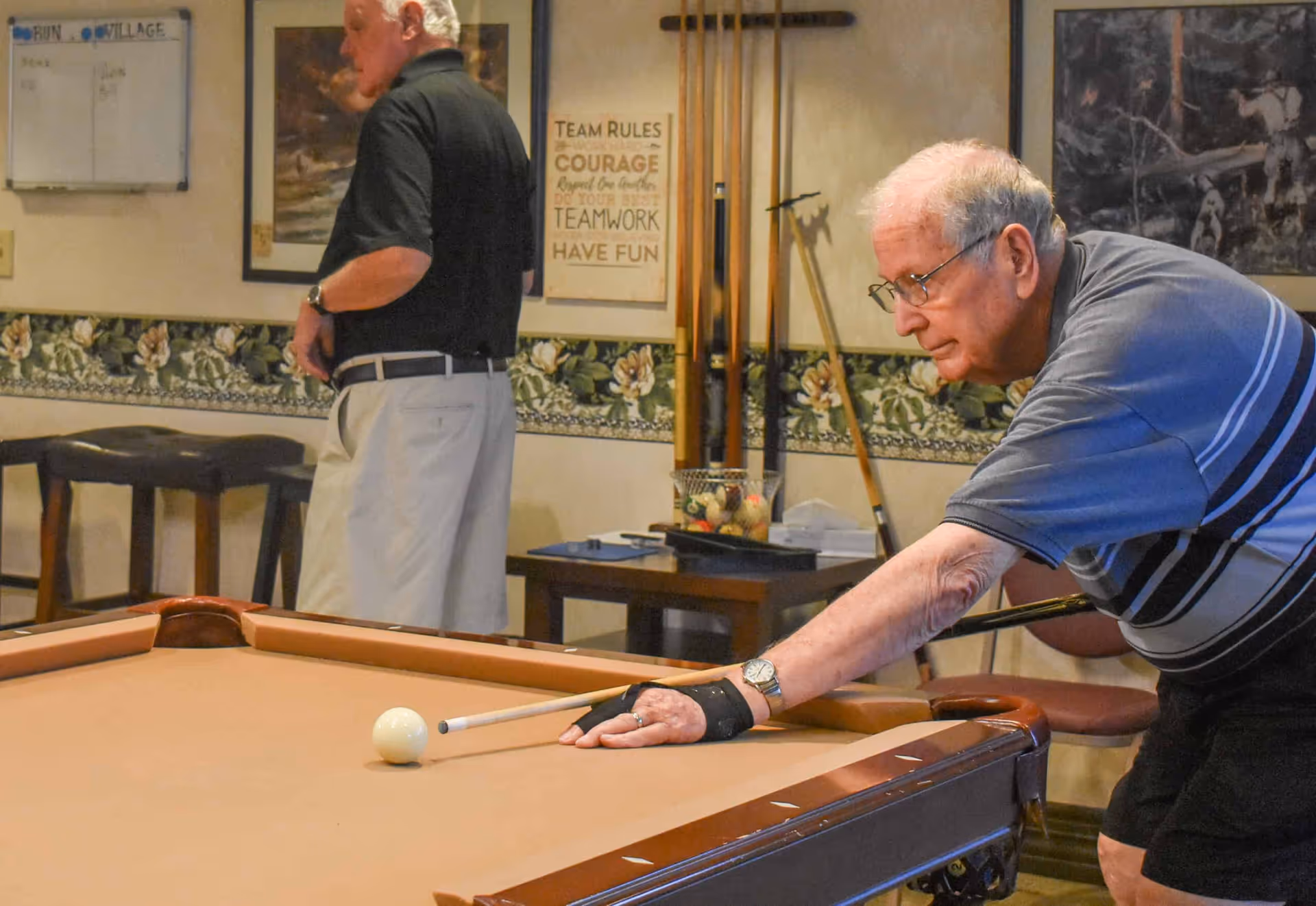 Two elderly men playing billiards in a recreational room with floral wallpaper and framed pictures on the walls. One man is aiming with a cue stick at the pool table while the other stands nearby.