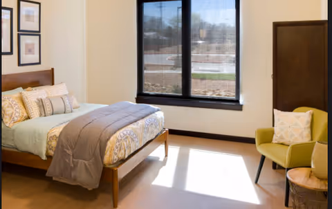 A bright bedroom with a wooden bed frame, neatly made bed with patterned bedding and multiple pillows, a green armchair with a decorative pillow, a small round wooden side table, and a large window with black trim letting in natural light.