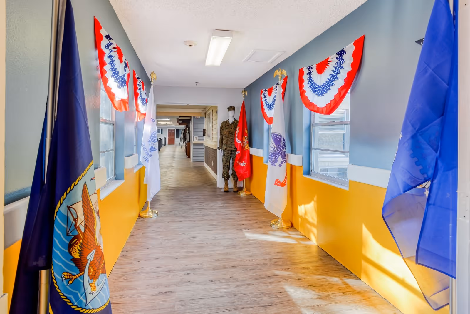 Hallway inside a senior living facility decorated with U.S. military flags and red-white-blue bunting, with a mannequin in uniform at the far end.
