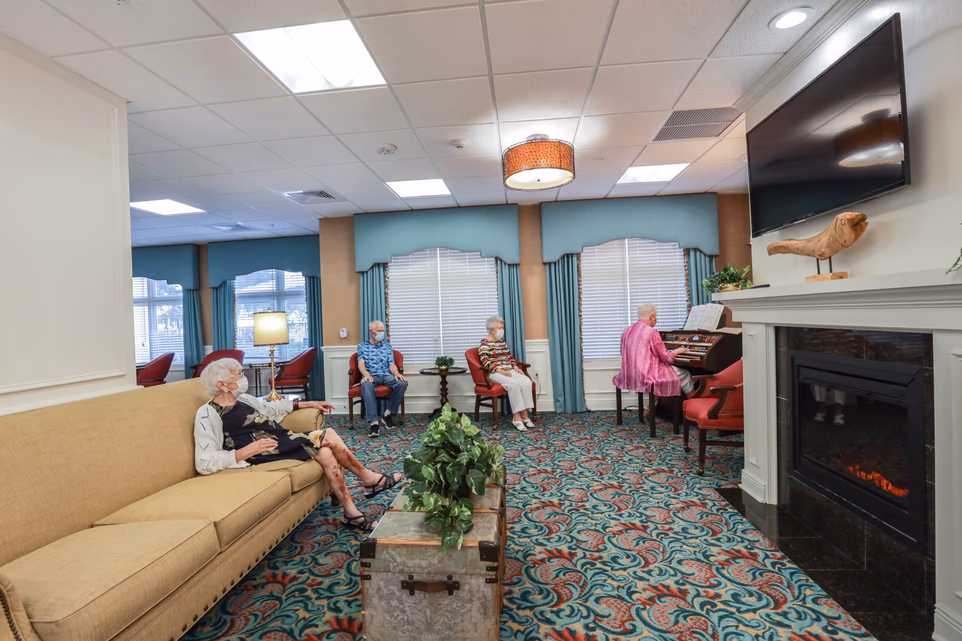A cozy living room in a senior living facility with patterned carpet, a beige sofa, and several elderly people seated. One person is playing an organ near a fireplace with a mounted flat-screen TV above it. The room has large windows with blue curtains and soft lighting.