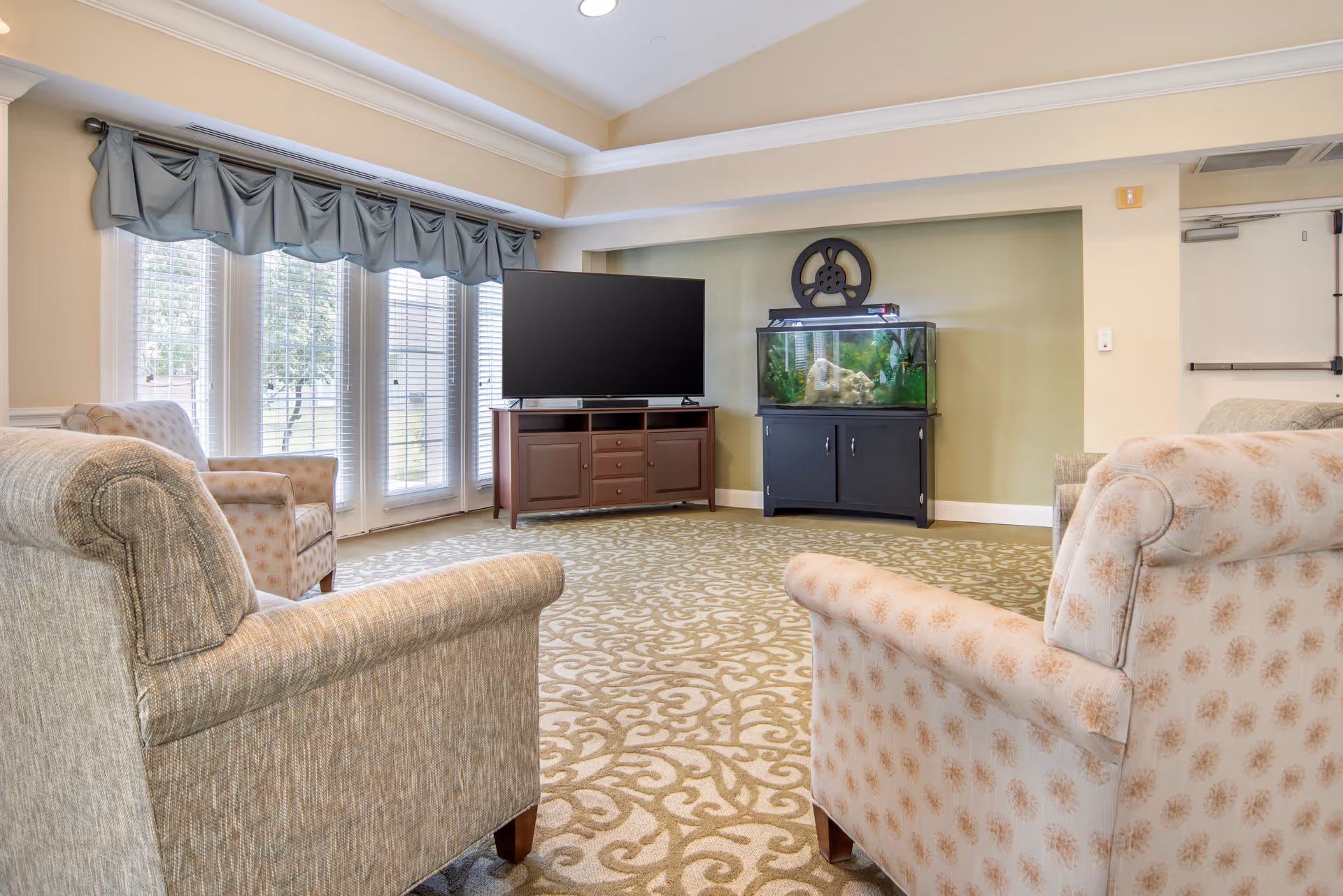 A cozy living room area with patterned carpet, three upholstered armchairs arranged around a flat-screen TV on a wooden stand, and a large fish tank on a black cabinet against a green accent wall. Large windows with blue valances allow natural light to fill the room.