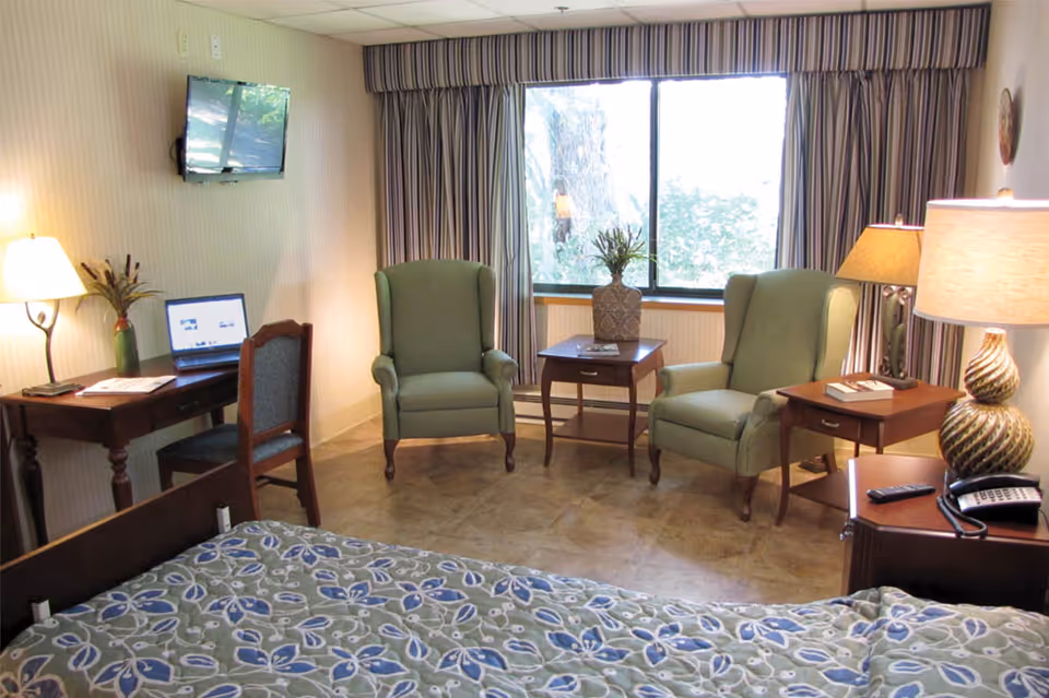 A cozy room with a bed covered in a floral patterned bedspread in the foreground. Two green upholstered armchairs are placed near a window with striped curtains. Between the chairs is a small wooden table with a decorative vase. There is a wooden desk with a chair, a laptop, and a lamp on the left side. A wall-mounted TV is above the desk. On the right side, a wooden nightstand holds a lamp, a phone, and a remote control.