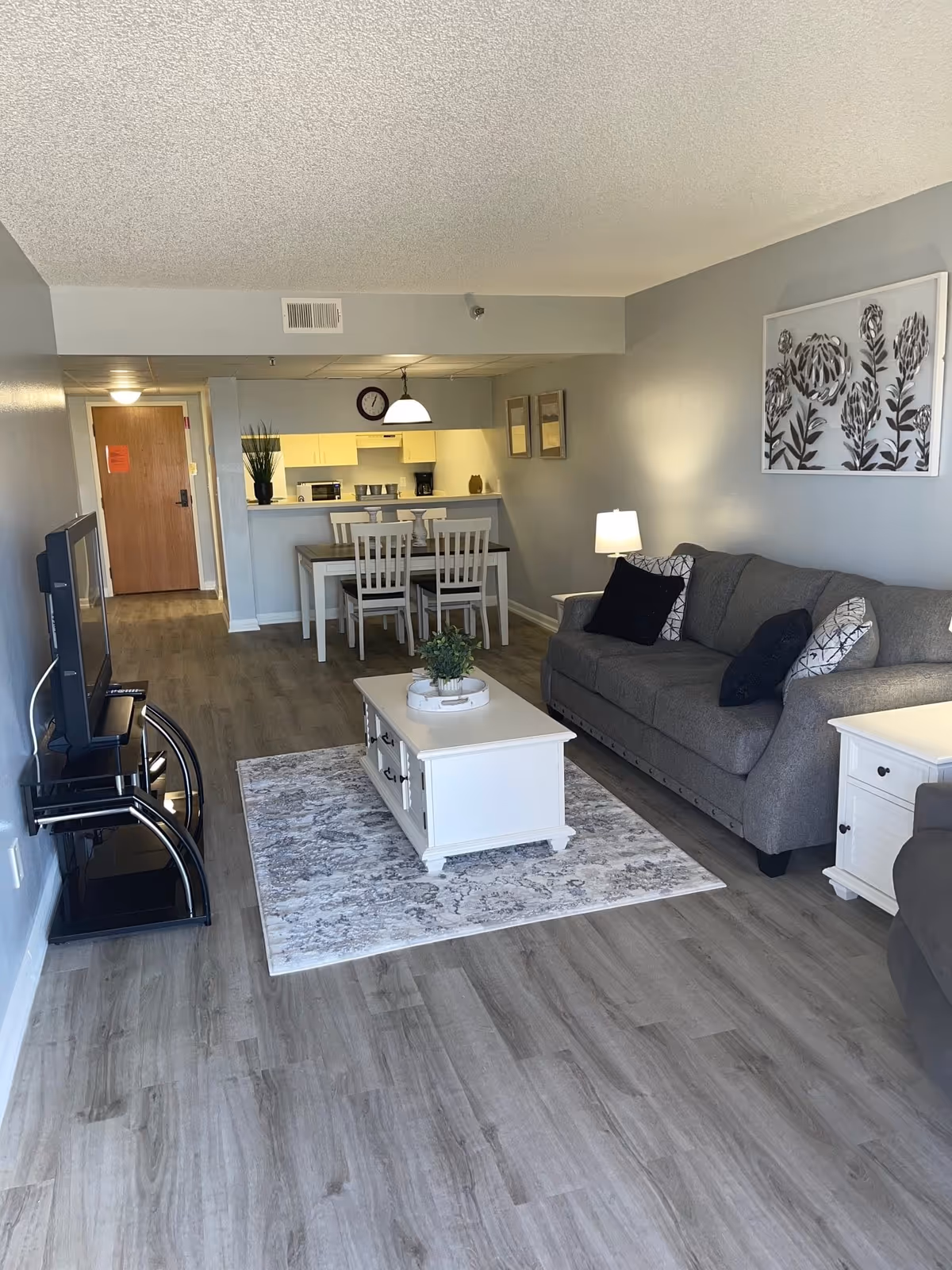 A cozy living room and dining area in a senior living facility. The room features a gray couch with patterned pillows, a white coffee table on a decorative rug, a TV on a black stand, and a dining table with four chairs. The walls are painted light gray with framed artwork and a lamp on a side table. The kitchen is visible in the background with white cabinets and appliances.