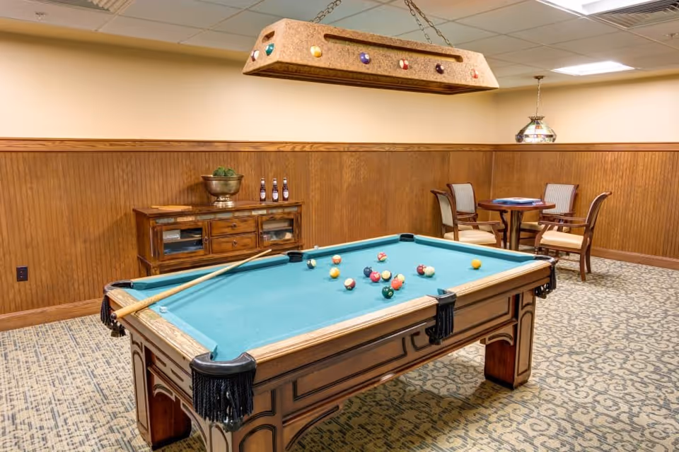 A recreational room with a pool table in the center, pool cues resting on the table, and billiard balls scattered on the green felt. The room has wood-paneled walls, a patterned carpet, a wooden cabinet with decorative items and bottles, and a small round table with four chairs in the corner. A hanging light fixture with billiard ball designs is above the pool table.