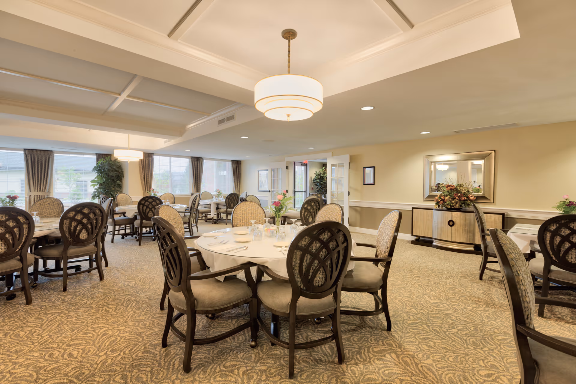 A spacious dining room with multiple round tables covered with white tablecloths, each set with plates, glasses, and silverware. The room features patterned carpet, cushioned chairs with dark wooden frames, large windows with curtains, and a sideboard with a decorative floral arrangement under a large mirror. Soft ceiling lights illuminate the area.