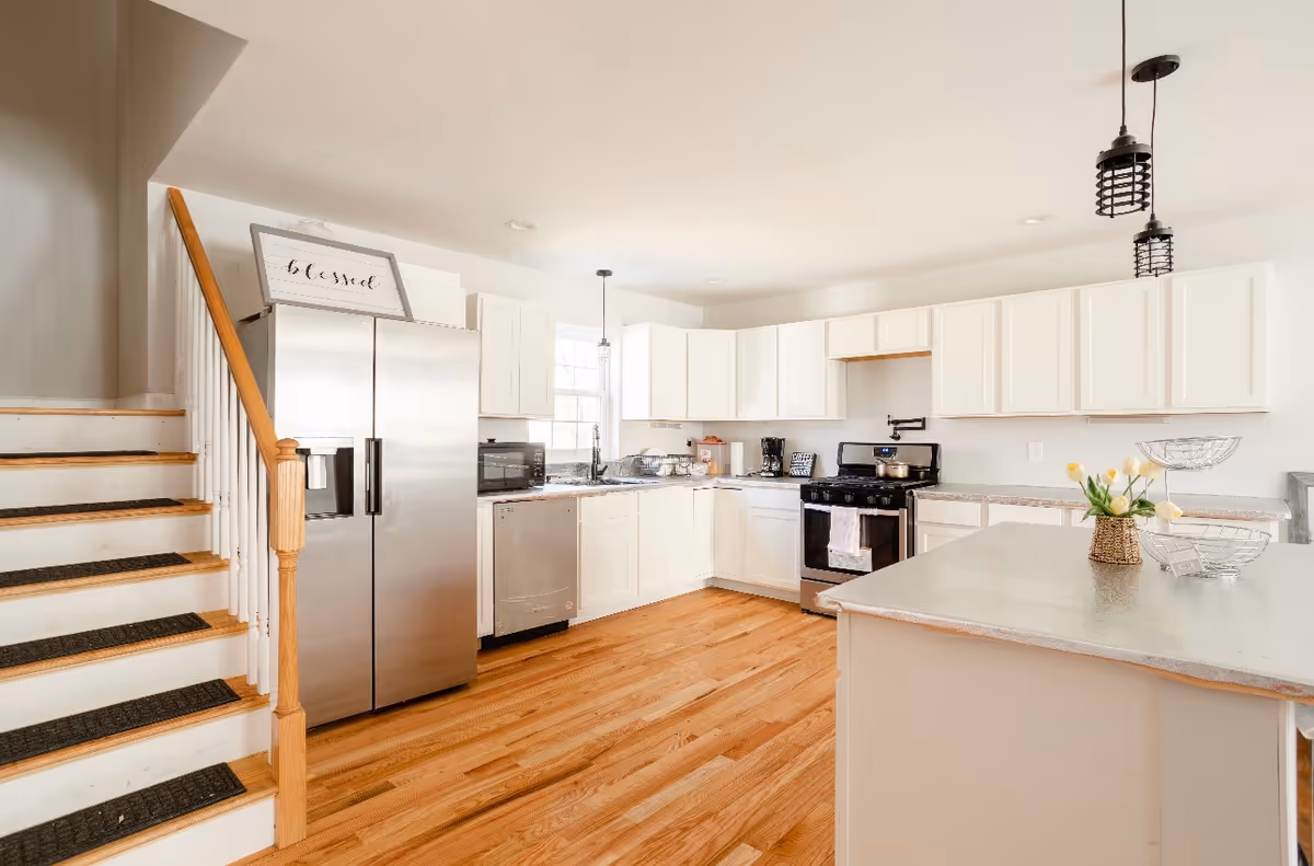A bright and modern kitchen with white cabinets, stainless steel refrigerator, dishwasher, and stove. There is a kitchen island with a vase of yellow tulips and a wire fruit basket. Wooden stairs with black stair treads are visible on the left side. The kitchen has wooden flooring and pendant lights hanging from the ceiling.