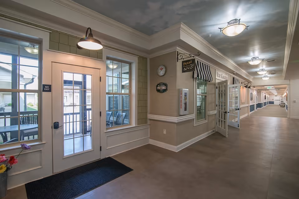 Interior hallway of The Lantern at Morning Pointe Alzheimer's Center of Excellence Chattanooga with a door labeled 'No Exit' leading to an outdoor patio area with chairs and tables. The hallway features beige walls, ceiling lights, a clock, and a small storefront window with a striped awning labeled 'Ice Cream'.