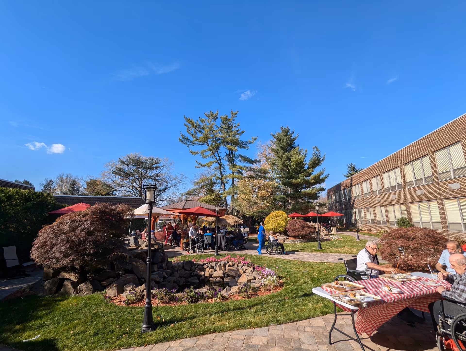 Outdoor courtyard area at Regency Jewish Heritage Nursing and Post-Acute Rehabilitation Center with people sitting and socializing under red umbrellas. There are trees, bushes, a small rock garden with flowers, and a brick building in the background under a clear blue sky.