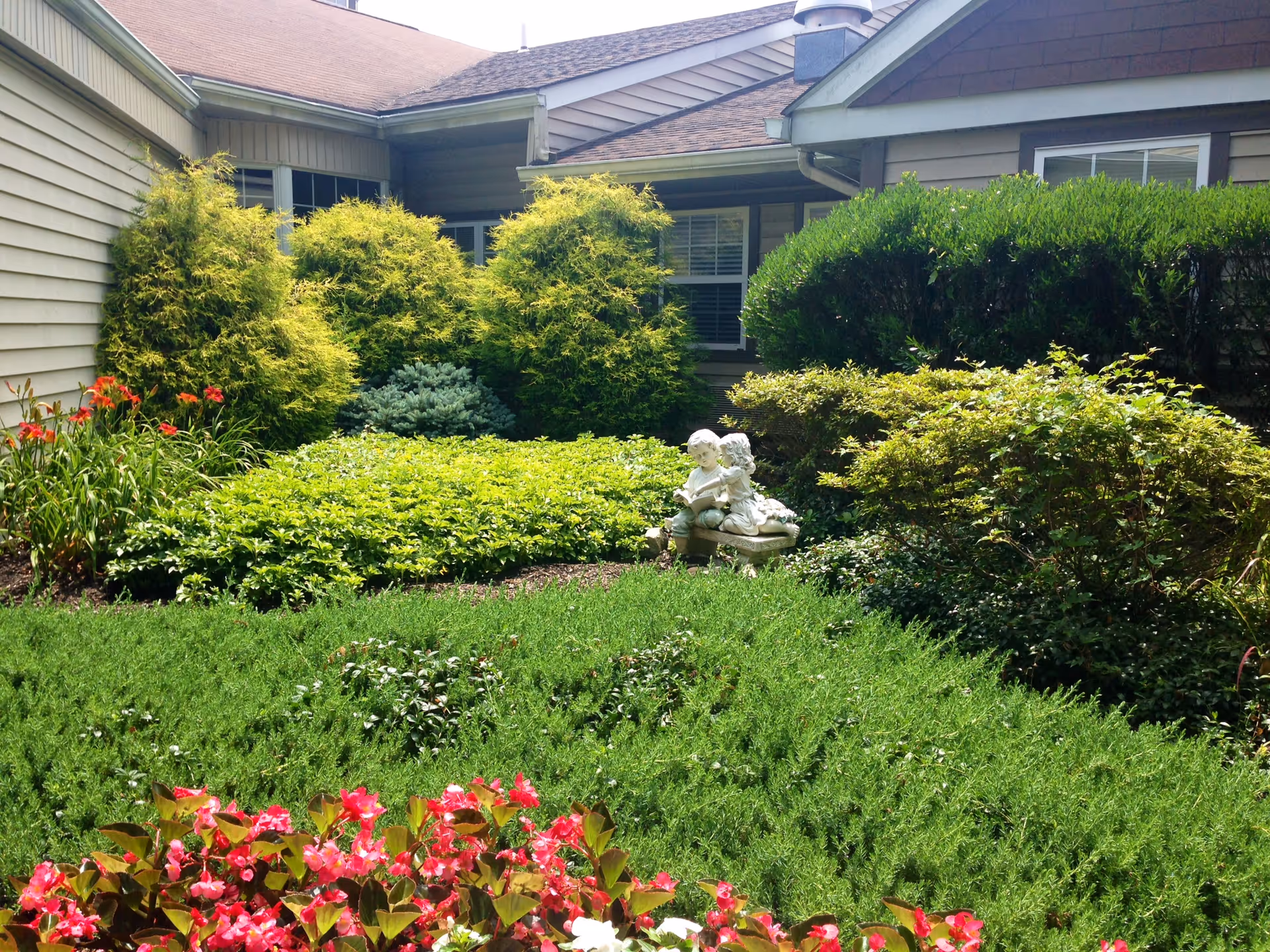 Sunny courtyard garden with shrubs, flowering plants, and a stone statue beside the building.