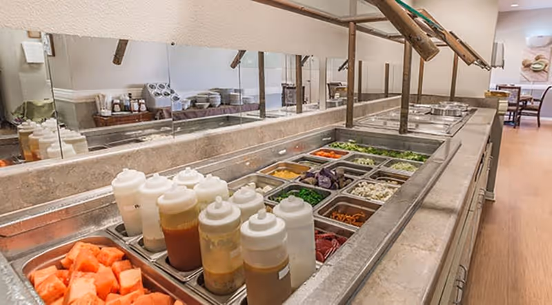 A salad bar with various fresh vegetables, toppings, and several squeeze bottles of dressings in a senior living facility dining area. The salad bar is set against a mirrored wall with a dining area visible in the background.