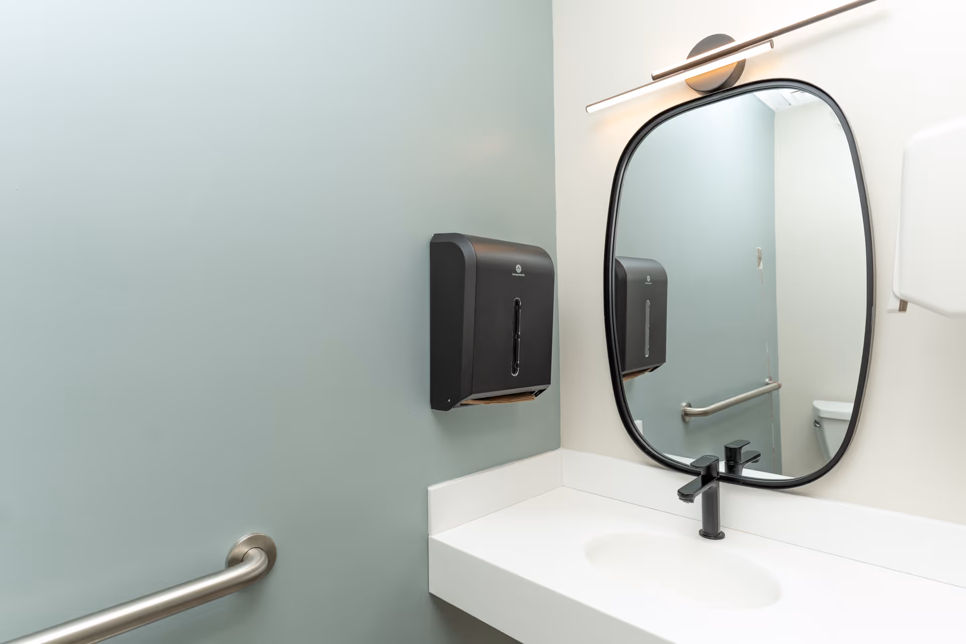 Accessible bathroom sink area with an oval mirror, black faucet, paper-towel dispenser and stainless steel grab bars against pale green and white walls.