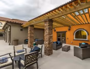 Outdoor patio area at HarborChase of Palm Coast featuring cushioned seating arrangements with chairs and sofas under a wooden pergola, stone pillars, and a yellow exterior wall with an arched window.