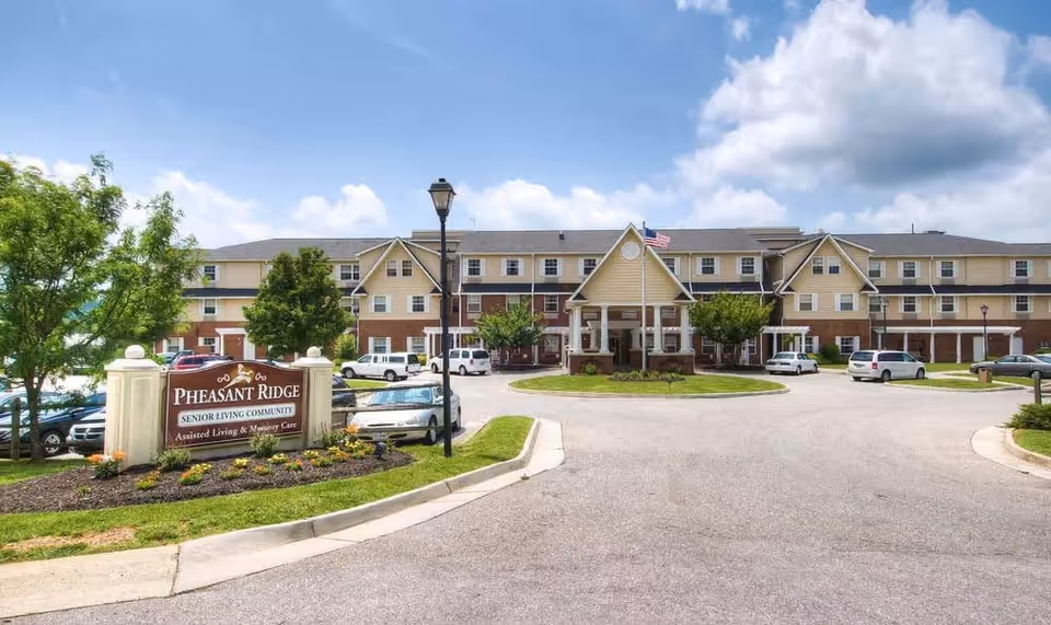 Front exterior of the Pheasant Ridge senior living community showing the building entrance with a porte-cochere, parking area, and a landscaped sign.